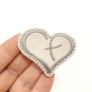 May include: White glitter heart-shaped patch with a silver embroidered cross in the center. The patch has a silver stitched outline.