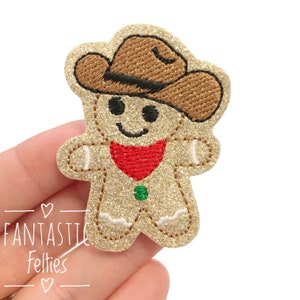 May include: A sparkly gold gingerbread man wearing a brown cowboy hat and a red bandana. The gingerbread man has a green button and a smiling face.