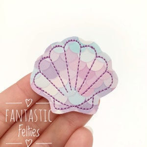 May include: A seashell-shaped felt applique with a purple stitched outline. The interior features a scale pattern in shades of purple, blue, and white. The text "FANTASTIC Felties" is visible in the lower left corner.