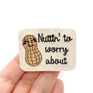 May include: White felt patch with a brown peanut cartoon with a smiling face and glasses. The text "Nuttin' to worry about" is embroidered in black.