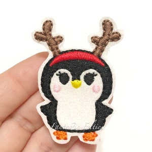 May include: A white felt penguin with black and white embroidered details, wearing a red headband and brown antlers. The penguin has a yellow beak and orange feet.