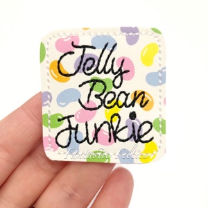 May include: A white felt patch with a colorful jelly bean pattern. The patch has black embroidery that reads "Jelly Bean Junkie" and "Fantastic Felties".