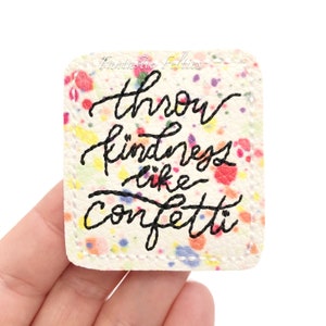 May include: White square patch with a colorful confetti pattern. The patch has black embroidered text that reads "Throw kindness like confetti."