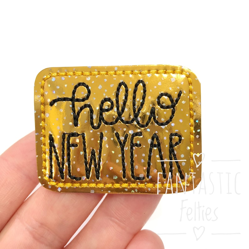 Puede incluir: Parche de planchado de purpurina dorada con texto bordado en negro que dice "hello new year".