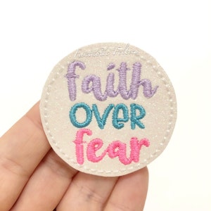 May include: A round, off-white felt patch with the embroidered words "faith OVER fear" in purple, teal, and pink. The patch has a glittery texture and a stitched border. The text is in a cursive font. The words are stacked vertically.
