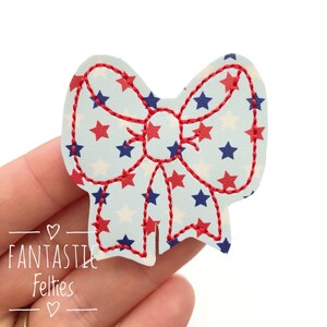 May include: A red and blue bow with white stars on a light blue background. The bow is made of felt and is perfect for adding a touch of patriotism to any project.