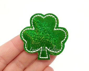 Puede incluir: Trébol verde con purpurina y costuras blancas. El trébol es un símbolo popular de la cultura irlandesa y se utiliza a menudo en decoraciones para el Día de San Patricio.