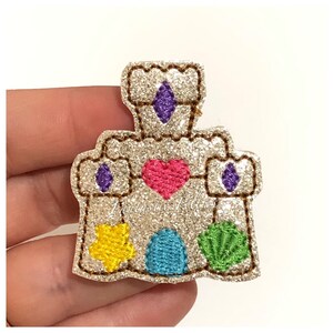 May include: Glittery castle applique with a heart, star, and seashell design. The castle features purple diamond shapes and a turquoise door. The applique is made of glitter fabric and has a brown stitched outline.