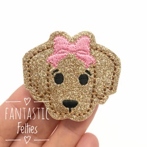 May include: A glittery gold felt dog head with a pink bow. The dog has black eyes and a black nose. The felt is stitched with brown thread. The text "FANTASTIC Felties" is below the dog.