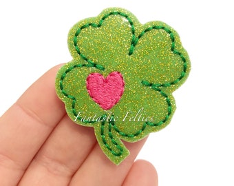 Puede incluir: Trébol verde con purpurina con un corazón rosa en el centro. El trébol está bordeado con hilo verde.