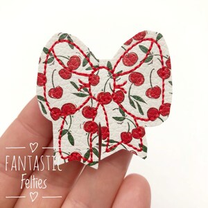 May include: A bow-shaped felt applique with a cherry pattern. The white felt is covered in red cherries and green leaves. Red thread outlines the bow and the cherry stems. The text "FANTASTIC Felties" is printed on the bottom left.