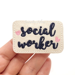 May include: A rectangular, glittery beige patch embroidered with the words "social worker" in navy blue script, with a small pink heart on each side. The patch has a stitched border and the text "Fantastic Felties" is visible.