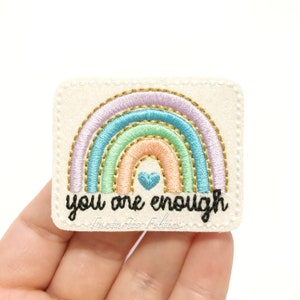 May include: A white felt patch with a rainbow embroidered in pastel colors and gold thread. The rainbow has a small blue heart in the center. The text "you are enough" is embroidered in black below the rainbow. The text "Fantastic Felties" is embroidered in black below the text.