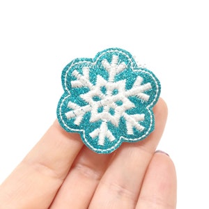 May include: A teal blue glitter snowflake applique with white embroidery. The snowflake is a 7-pointed star shape.