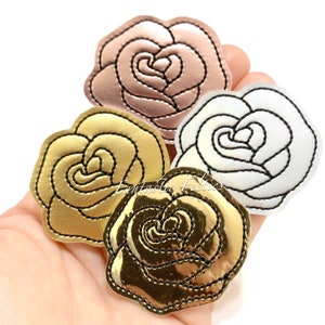 May include: Four rose-shaped felt appliques in metallic gold, rose gold, and white. Each applique features black stitching outlining the petals. The appliques are held in a hand, showcasing the different colors and textures.