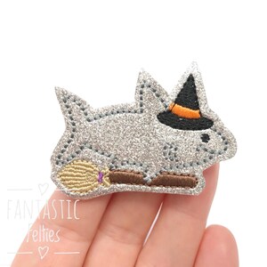 May include: A silver glitter felt applique in the shape of a stylized animal, wearing a black witch hat with an orange band. It holds a purple and brown broom. The text "FANTASTIC Felties" is visible.
