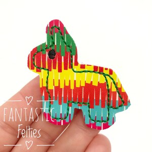 May include: A felt pinata applique in the shape of a traditional pinata. The pinata is decorated with red, yellow, green, and blue stripes and has a black eye.