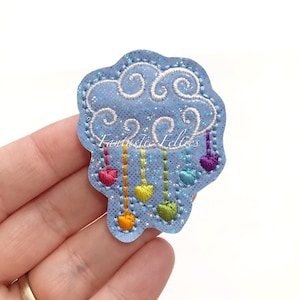 May include: A blue felt cloud with rainbow hearts hanging from it. The cloud has a sparkly finish and the hearts are embroidered with white stitching. The text "Fantastic Felties" is visible in the image.