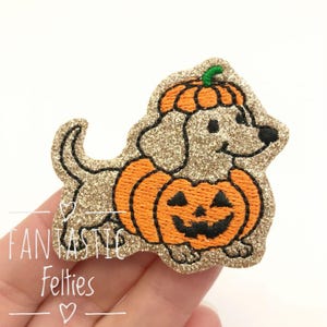 May include: A glittery gold embroidered patch shaped like a dachshund wearing a carved orange pumpkin costume with a green stem on its head. The pumpkin features a black jack-o'-lantern face. The text FANTASTIC Felties is visible below.