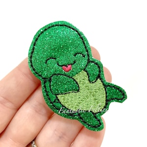 May include: A green glittery turtle-shaped felt applique with a smiling face and a red tongue sticking out. The turtle has a light green belly and black stitched outlines. The text "Fantastic Felties" is visible.