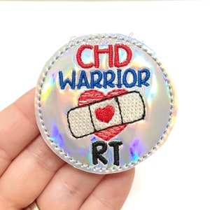 May include: A silver iridescent round patch with the text "CHD WARRIOR RT" in blue, red, and black embroidery. A red heart with a white bandage is in the center of the patch.