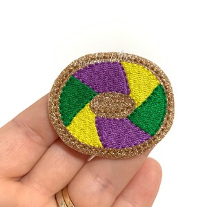 May include: A round, gold glitter-covered patch with a purple, green, and yellow swirl design. The patch is embroidered with a gold thread.