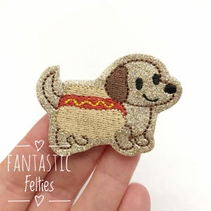 May include: Glittery gold felt dog applique with brown ears and a hot dog body. The hot dog has a red and yellow zigzag pattern. The applique has the text "Fantastic Felties".