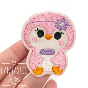May include: Embroidered penguin patch in pink, white, and purple with glitter accents. The penguin has a purple flower on its head and orange feet. The text "Fantastic Felties" is visible in the lower left corner.