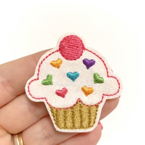May include: A white cupcake with a pink cherry on top and rainbow hearts embroidered on the frosting. The cupcake is on a white background.