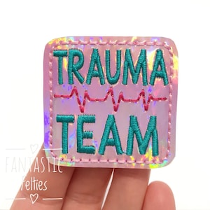 May include: A pink iridescent iron-on patch with the text "Trauma Team" in teal embroidery. A red embroidered EKG line is above the word "Team".