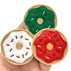May include: Three felt donut ornaments in green, red, and white with brown, white, and green sprinkles. The donuts are on a hand.