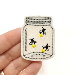 May include: A white mason jar with a gray lid filled with three glowing fireflies. The jar is on a white background with the text "Fantastic Follies" at the bottom.
