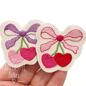 May include: Two felt applique patches with embroidered cherries and bows. One patch has a pink bow and the other has a purple bow. Both patches have green stems and red and pink cherries.