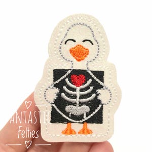 May include: A white felt applique in the shape of a smiling cartoon bird, with an orange beak and feet. The bird has a black and white skeleton design on its chest, with a red heart. The text "ANTASITC Felties" is on the left side.