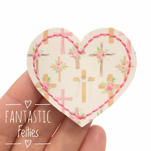 May include: A heart-shaped felt applique with a pink stitched border. The applique features a pattern of crosses in shades of pink and gold, some adorned with floral bouquets. The text "FANTASTIC Felties" is visible.