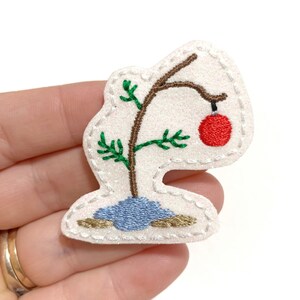 May include: A felt Christmas tree ornament with a single brown branch, green needles, and a red ornament. The tree is sitting on a blue base with brown rocks.
