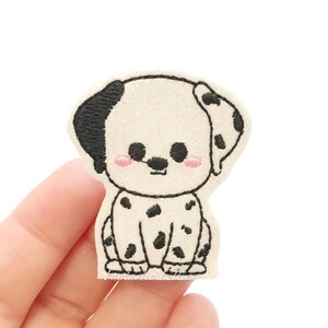 May include: White and black embroidered felt patch of a cartoon Dalmatian dog with pink cheeks. The dog is sitting with its legs crossed and has a black nose and eyes.