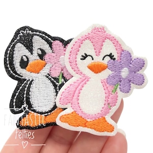 May include: Two embroidered penguin patches with glittery fabric. One penguin is black and white holding a pink flower, and the other is pink and white holding a purple flower. Both have orange beaks and feet. The text Fantastic Felties is visible.