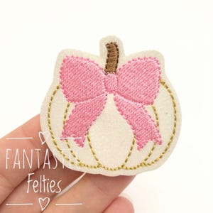 May include: An embroidered pumpkin patch featuring a white glittery base, a brown stem, and a large pink bow centered on the front. The pumpkin is outlined with gold stitching. The text FANTASTIC Felties is visible in the lower left corner.