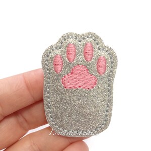May include: A gray, glittery paw print patch with pink embroidered paw pads. The patch has a stitched border and is likely an iron-on or sew-on applique. The text "Fantastic Feltries" is visible at the bottom.