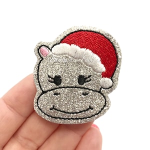 May include: A silver glittery embroidered patch shaped like a hippo head wearing a red and white Santa hat. The hippo has black embroidered eyes with eyelashes, a pink inner ear, and a stitched smile. The text Fantastic Felties is visible.