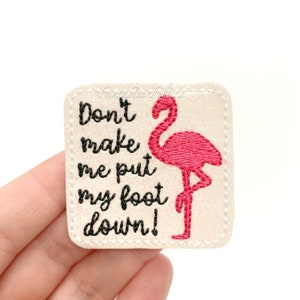 May include: White iron-on patch with pink glitter and black embroidery. The patch features a pink flamingo and the text "Don't make me put my foot down!"