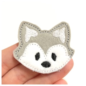May include: Gray felt applique of a wolf face with white ears, nose, and eyes. The applique is embroidered with black stitching.