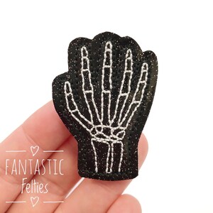 May include: A black glittery patch in the shape of a hand, featuring a white embroidered skeleton hand design. The patch has the text "Fantastic Felties" in pink.