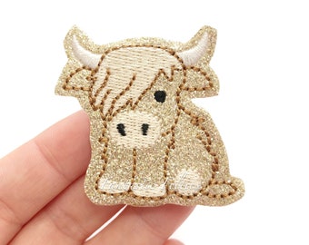 May include: A gold glitter embroidered patch of a cartoon Highland cow with black eyes and a white nose. The patch is about 2 inches tall and 2 inches wide.