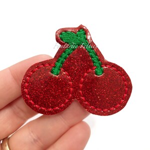 May include: Two red glitter cherries with green stems and leaves. The cherries are sewn together and have a red stitched outline.