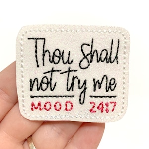 May include: A rectangular, off-white felt patch with the text "Thou shalt not try me" in black script. Below, in red, are the words "MOOD 24/7". The patch has a glittery texture and a stitched border.