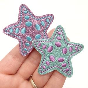 May include: Two glitter star-shaped fabric patches with embroidered details. One is pink with blue dots and the other is blue with pink dots.