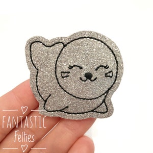 May include: A glittery silver seal-shaped felt applique with a black outline. The seal has a smiling face with closed eyes and a small black nose. The text "Fantastic Felties" is printed in white.