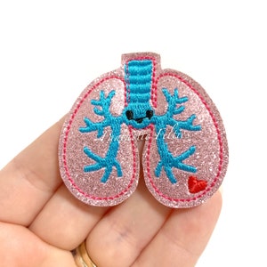 May include: Pink glitter felt applique of a cartoon lung with a blue airway and a red heart. The lung has a smiling face.